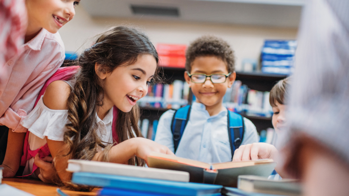 A diverse group of elementary school students look at a book together, smiling.