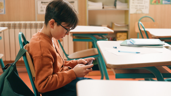 A boy with glasses sits at a school desk looking down at a cellphone.