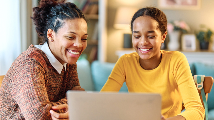 A Black mother and daughter sit at a table, smiling while looking at a computer.